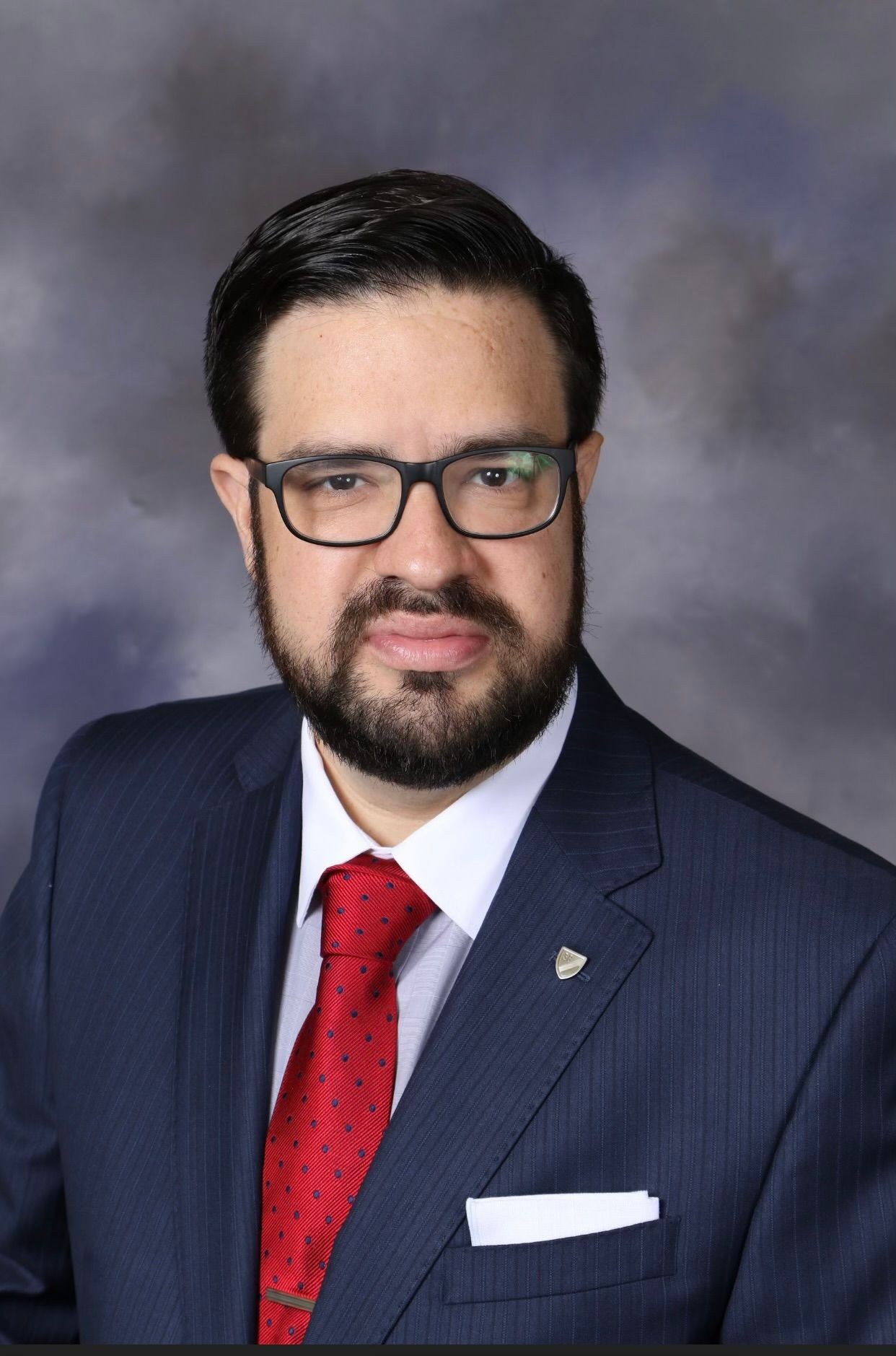 Man with glasses in a suit and red tie, posing against a gray background.