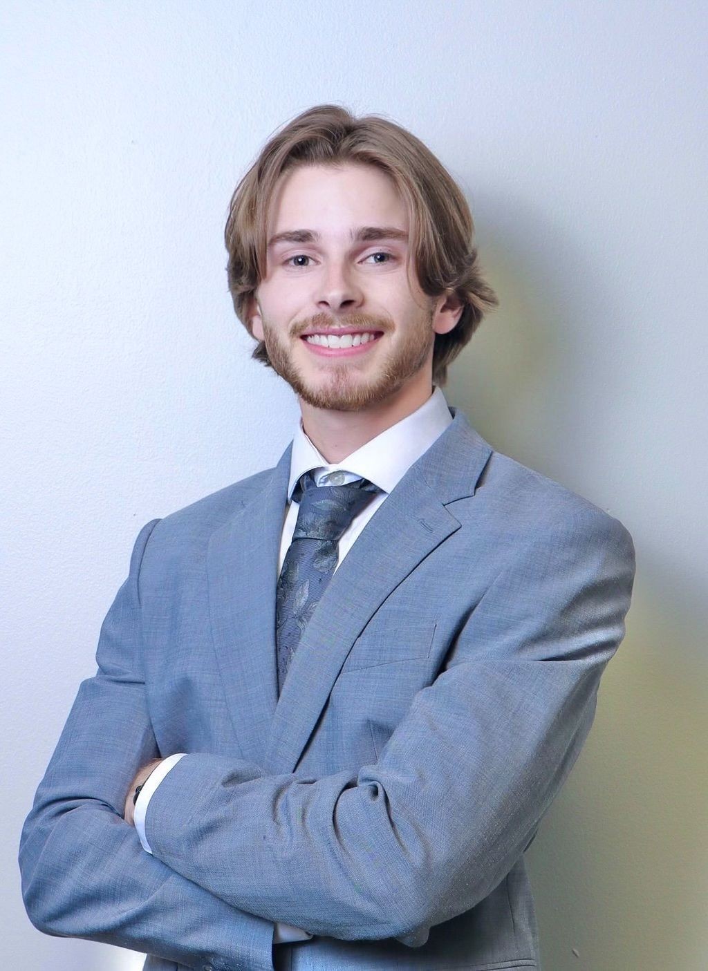 Person in a light blue suit and patterned tie standing with folded arms against a plain wall.