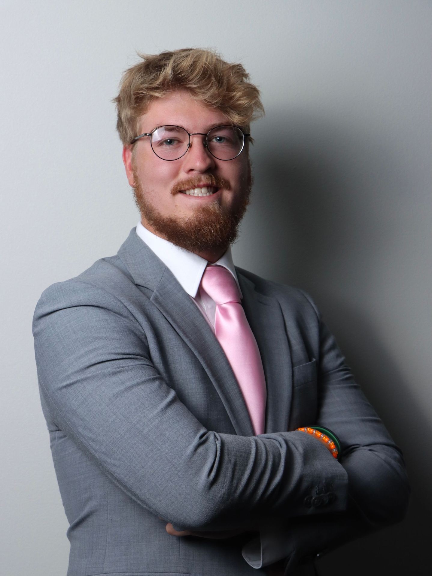 Man in glasses and gray suit with pink tie smiling, standing with arms crossed.