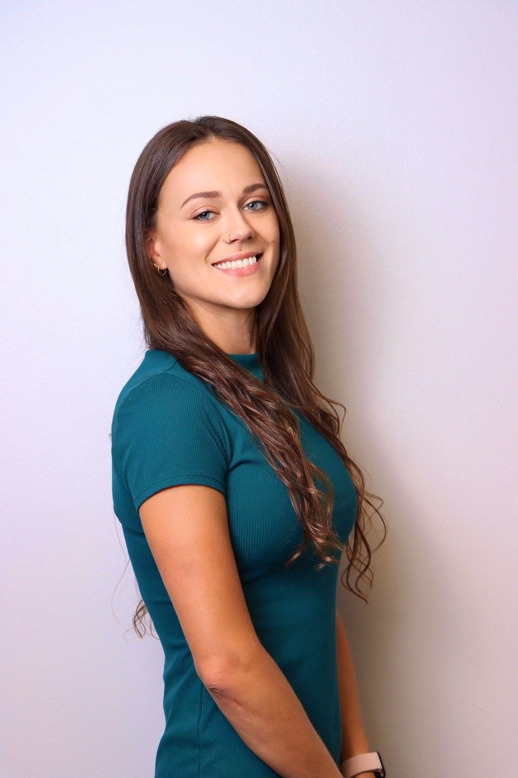 Woman with long brown hair smiling, wearing a teal shirt, against a plain background.